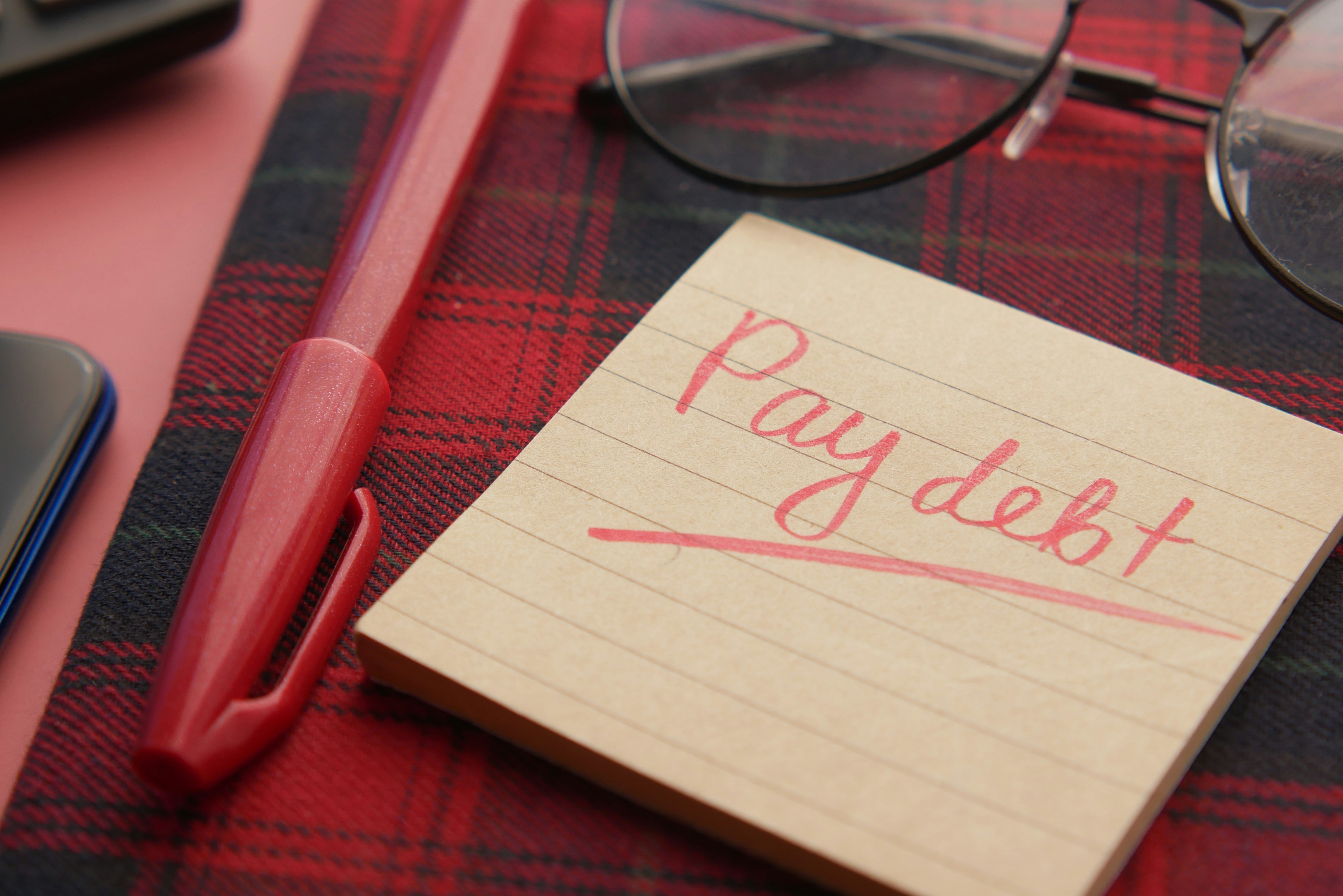 Close-up of a handwritten note reading “Pay debt” beside a pen and glasses, representing debtor tracing and debt recovery investigations.